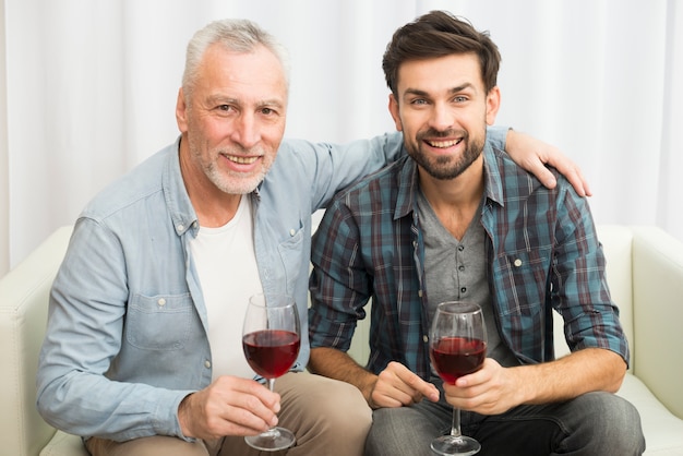 Uomo invecchiato che abbraccia il giovane ragazzo sorridente con bicchieri di vino sul divano
