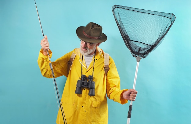 Uomo invecchiato con un impermeabile giallo e occhiali sta andando a pescare. Un uomo con una canna da pesca e una rete, parete isolata