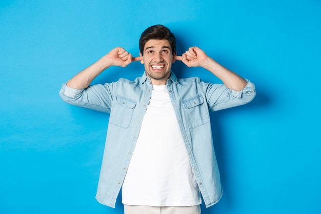 Uomo irritato chiudere le orecchie dai vicini rumorosi, alzando lo sguardo e facendo smorfie, ascoltando musica ad alto volume, in piedi sopra il muro blu