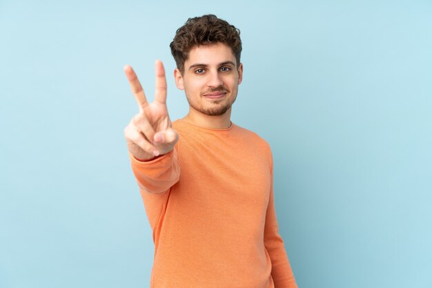 Uomo isolato sulla parete blu che sorride e che mostra il segno di vittoria