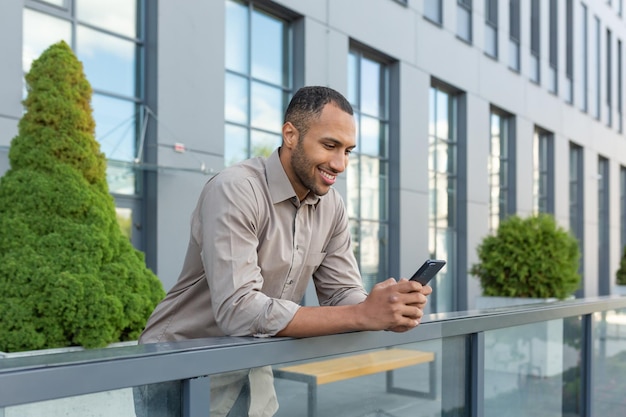 Uomo ispanico al di fuori di un moderno edificio per uffici utilizzando smartphone uomo d'affari in camicia digitando il messaggio e