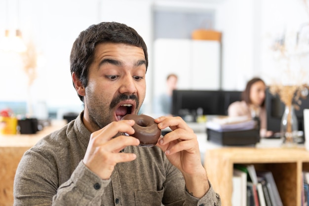 Uomo ispanico con baffi che mangia un panino dolce al cioccolato Abitudini malsane nel concetto di ufficio