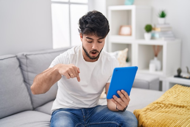 Uomo ispanico con barba che usa il touchpad seduto sul divano rivolto verso il basso con le dita che mostrano pubblicità, faccia sorpresa e bocca aperta