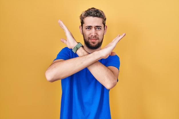 Uomo ispanico con barba in piedi su sfondo giallo espressione di rifiuto che incrocia le braccia facendo segno negativo, faccia arrabbiata