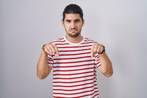 Uomo ispanico con capelli lunghi in piedi su sfondo isolato rivolto verso il basso guardando triste e sconvolto, indicando la direzione con le dita, infelice e depresso.