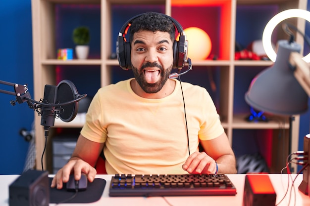 Uomo ispanico con la barba che gioca ai videogiochi con le cuffie che sporgono la lingua felice con un'espressione divertente. concetto di emozione.