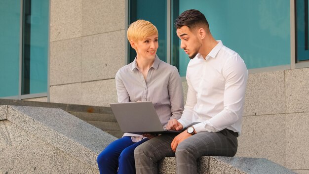 uomo ispanico e donna caucasica in piedi vicino al centro business in strada e lavorando al computer portatile