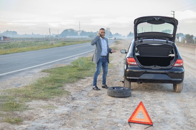 Uomo latino che chiama al cellulare accanto alla sua auto con una gomma a terra