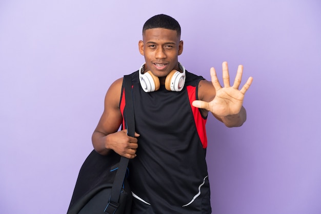 Uomo latino di giovane sport con borsa sportiva isolato su viola contando cinque con le dita