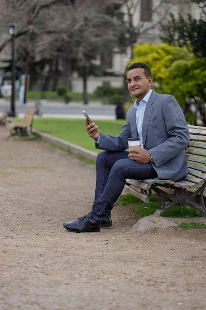 Uomo latino in giacca con una tazza di caffè usa e getta seduto su una panchina in un parco pubblico