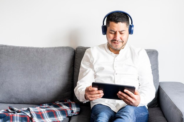 Uomo latinoamericano che guarda sul tablet un film o una serie con le sue cuffie wireless nel comfort del divano di casa