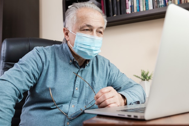 Uomo maggiore in maschera facciale che lavora o comunica sul computer portatile a casa. studio, formazione, lavoro, comunicazione, intrattenimento, tempo libero durante il periodo del coronavirus.