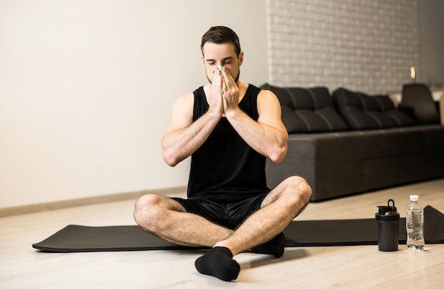 Uomo malato con naso che cola seduto sul tappetino yoga nero. Resta a casa concetto. Il giovane castana si soffia il naso dopo l'allenamento. Praticare sport a casa se ti senti male.