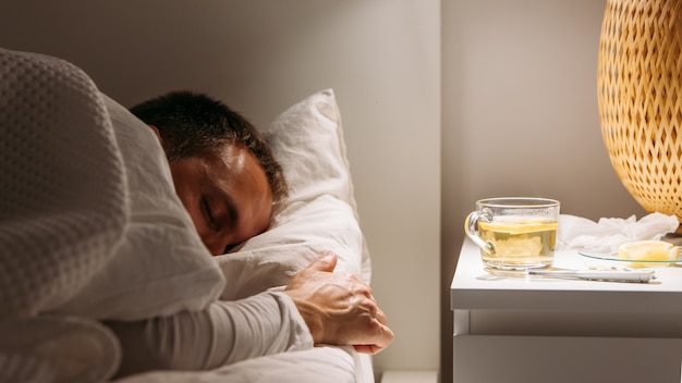 Uomo malato dorme nel letto con la febbre alta, affetto da influenza, tazza di tè al limone sul tavolo