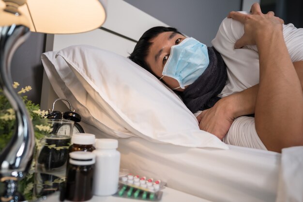 Uomo malato nella maschera medica che ritiene freddo e che soffre dalla malattia del virus e dalla febbre a letto, concetto di pandemia del coronavirus.