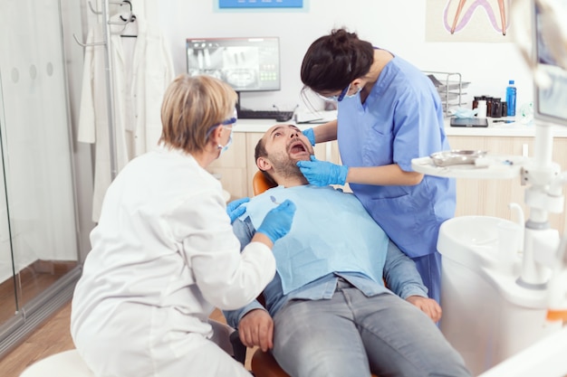 Uomo malato seduto sulla poltrona del dentista con la bocca aperta mentre infermiere medico con maschera facciale e guanti che analizzano la salute dei denti durante la consultazione stomatologia