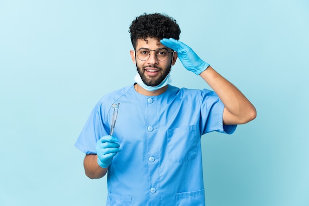 Uomo marocchino del dentista che tiene gli strumenti isolati sull'azzurro con l'espressione di sorpresa