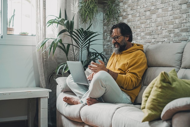 Uomo maturo a casa seduto sul divano a fare videochiamate con il laptop Uomini felici e moderni lavorano su un laptop e connessione internet wireless Persona intelligente che lavora al chiuso Tecnologia e tempo libero