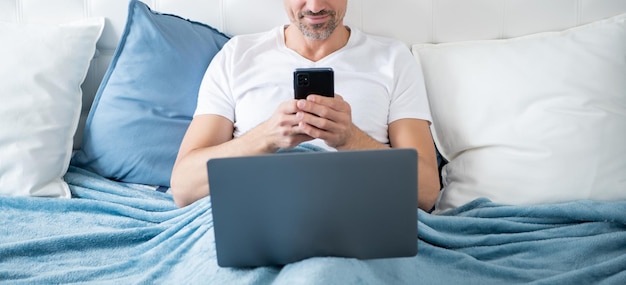 Uomo maturo allegro che chiacchiera sul telefono e lavora al computer portatile a letto