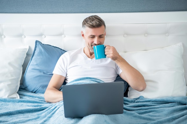 Uomo maturo allegro che lavora al computer portatile a letto con caffè