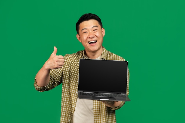 Uomo maturo asiatico felice che mostra lo schermo vuoto del computer portatile e che gesturing il pollice in su che posa sopra il verde