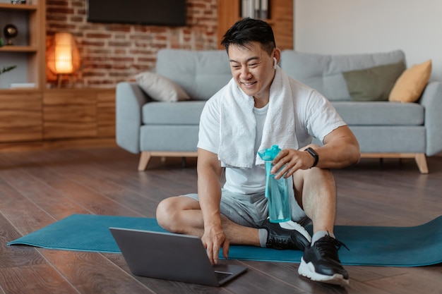 Uomo maturo asiatico sportivo felice che si siede sulla stuoia di yoga con la bottiglia d'acqua che sceglie video di forma fisica su Internet facendo uso del computer portatile