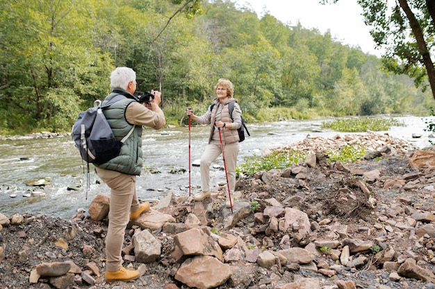 Uomo maturo attivo con zaino e macchina fotografica che fotografa la sua moglie felice con i bastoni da trekking sulla riva del fiume