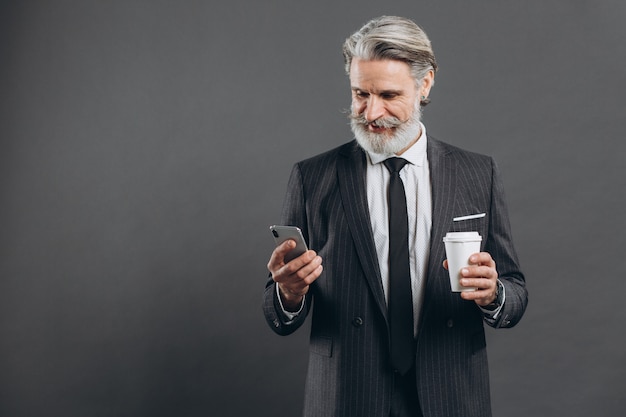 Uomo maturo barbuto alla moda e di affari in un vestito grigio che guarda sul telefono, bevendo caffè e sorridendo sulla parete grigia.