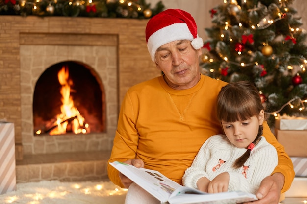 Uomo maturo che legge la storia con la sua nipote in soggiorno decorato con albero di Natale e ghirlande, bambino e nonno seduto vicino al camino e guardando attentamente le pagine del libro.