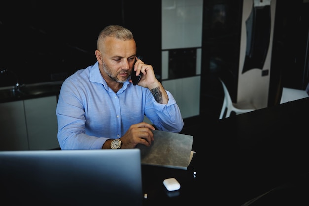 Uomo maturo che parla al telefono mentre lavora al laptop in remoto a casa