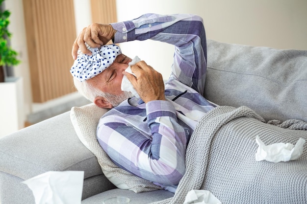 Uomo maturo che si sente male con il raffreddore e la febbre a casa malato di influenza seduto sul divano con un pacchetto di ghiaccio sulla testa