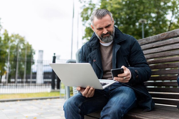 Uomo maturo con barba e capelli grigi lavora fuori con laptop e telefono