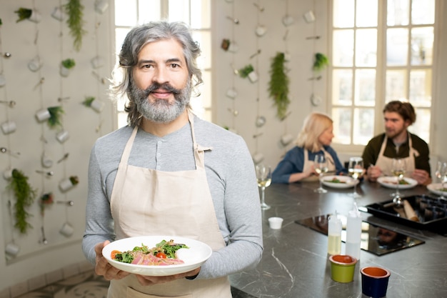 Uomo maturo con la barba in pullover e grembiule che tiene piatto con insalata di verdure e carne fatta in casa mentre si trova in piedi contro due persone che mangiano il pranzo