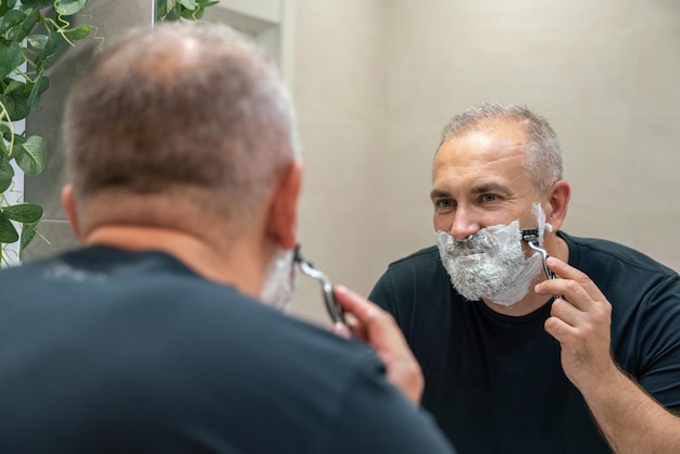 Uomo maturo dai capelli grigi che si rinnova la barba a casa usando il rasoio