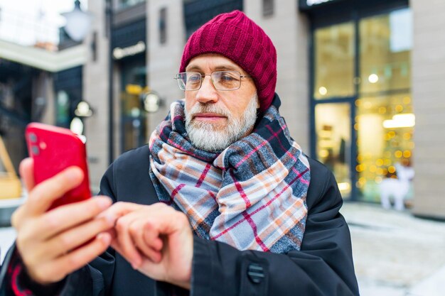 Uomo maturo di bell'aspetto con cappello e occhiali utilizzando l'app del telefono cellulare che invia messaggi all'esterno