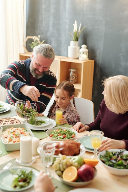 Uomo maturo felice che dà insalata alla sua adorabile nipote dalla tavola festiva durante la cena in famiglia in vacanza invernale