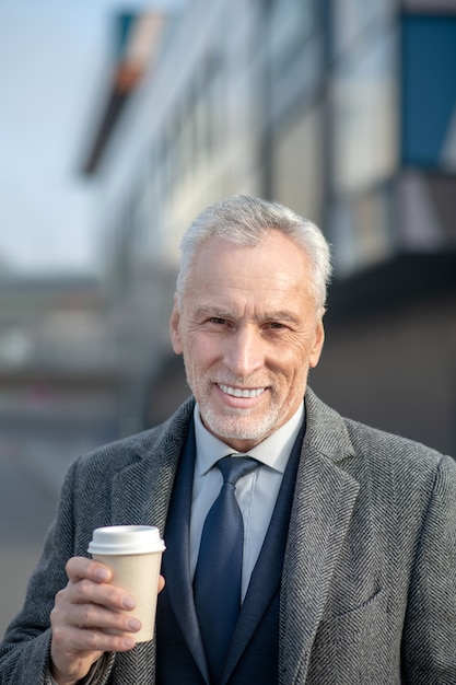 Uomo maturo in cappotto elegante che sorride piacevolmente mentre beve il caffè