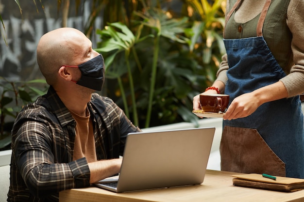 Uomo maturo in maschera protettiva che lavora al tavolo con il computer portatile mentre il cameriere che serve il caffè al ristorante