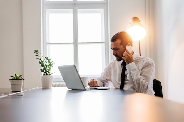 Uomo maturo occupato in camicia e cravatta lavorando al computer portatile e parlando al telefono