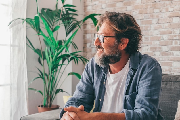 Uomo maturo premuroso che guarda fuori attraverso la finestra seduto sul divano in un appartamento di lusso moderno Contemplativo maschio caucasico sorridente che ammira la vista seduto comodamente sul divano di casa