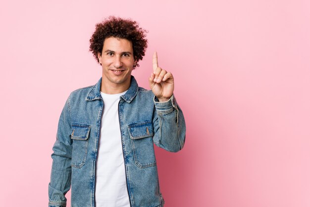 Uomo maturo riccio che porta una giacca di jeans contro la parete rosa che mostra numero uno con il dito.