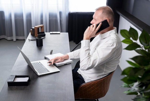 Uomo maturo seduto alla scrivania con laptop e pianificatore. Uomo d'affari che parla al telefono con un collega o un partner commerciale. Concetto di chiamata d'affari. Foto di alta qualità