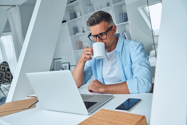 Uomo maturo sicuro che lavora al computer portatile e beve caffè mentre è seduto alla scrivania dell'ufficio