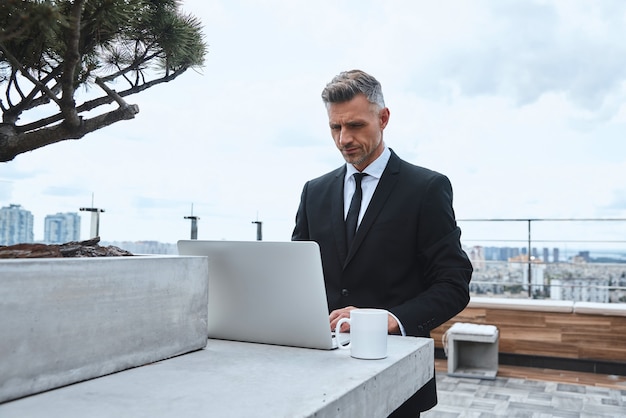 Uomo maturo sicuro che lavora al computer portatile mentre sta in piedi sulla terrazza sul tetto