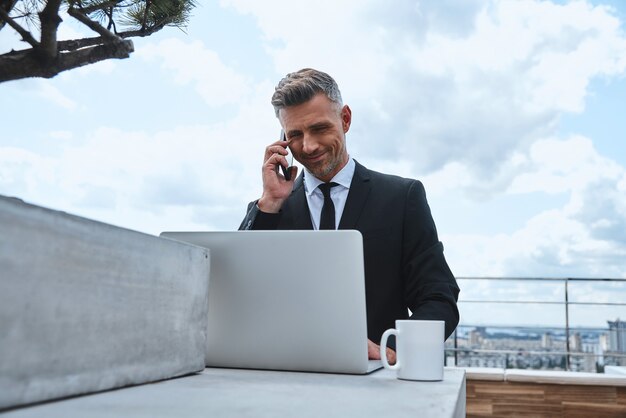 Uomo maturo sicuro che lavora al laptop e parla al cellulare mentre si trova sul ter...