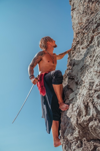 Uomo maturo tatuato che tiene la spada pronta a combattere e arrampicarsi sulla roccia con sfondo blu