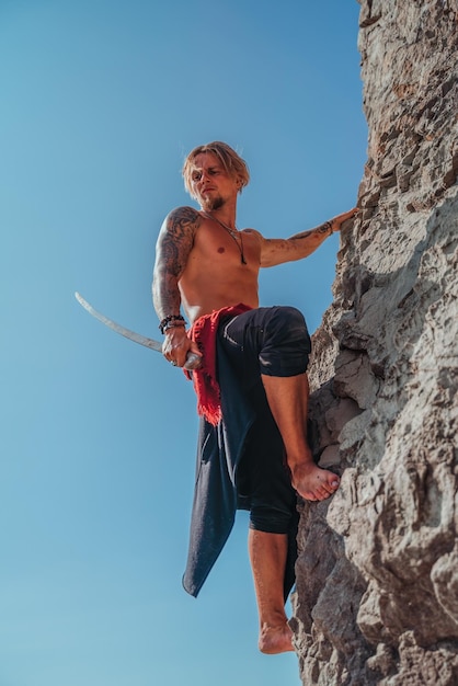 Uomo maturo tatuato che tiene la spada pronta a combattere e arrampicarsi sulla roccia con sfondo blu