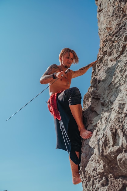 Uomo maturo tatuato che tiene la spada pronto a combattere e arrampicarsi sulla roccia con sfondo di cielo blu - Ritratto di un bel guerriero antico muscoloso con una spada. Concetto di arte marziale