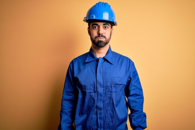 Uomo meccanico con barba che indossa uniforme blu e casco di sicurezza su sfondo giallo guance gonfie con faccia buffa Bocca gonfiata con aria espressione pazza