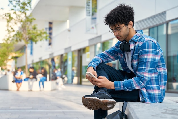 Uomo mediorientale pensieroso che utilizza messaggi di testo sul telefono cellulare per strada. Shopping online per hipster